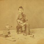 Little_boy_with_a_block_of_wood_carving_tools_and_a_doll_by_Lamprey_M._S._Maurice_S..png